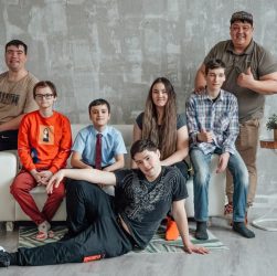 Group of seven people posing casually in a stylish indoor setting with a concrete wall background.