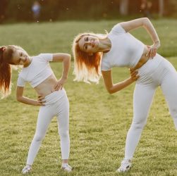 A mother and daughter in activewear stretching on a sunny day in a grassy field.
