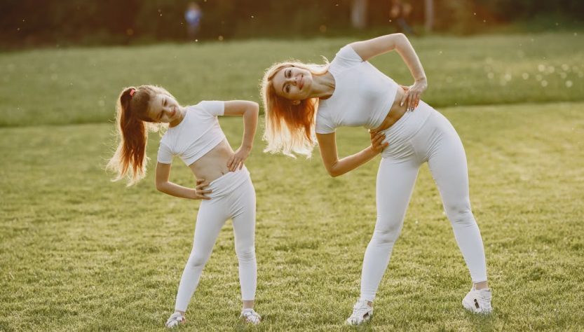 A mother and daughter in activewear stretching on a sunny day in a grassy field.