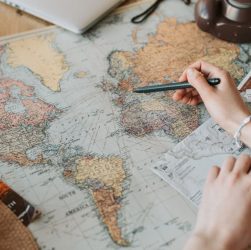 Hands marking destinations on a world map surrounded by travel essentials like passport and camera.