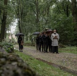 Mourners walk through a peaceful, wooded cemetery during a funeral procession.